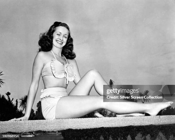 American actress Noel Neill sitting on a diving board in a two-piece, frilled bathing costume, circa 1945.