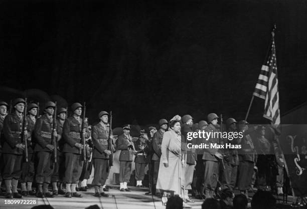 American soprano Dorothy Maynor singing on stage beside a formation of US Army soldiers and a raised American flag during a concert, circa 1950.