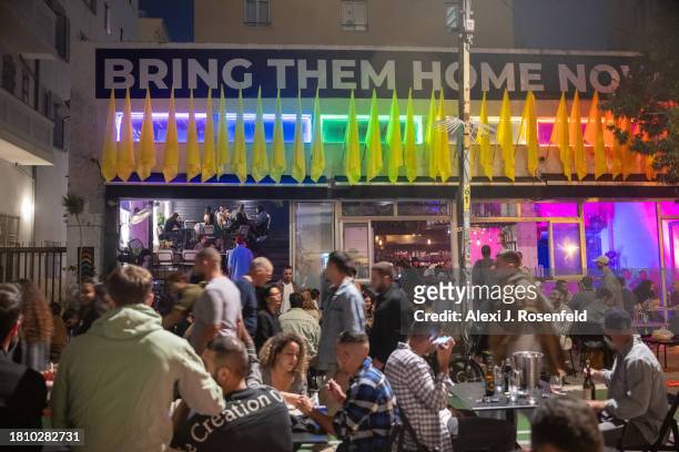 People sit at tables at the street dining and drinking at the bar Shpagat where a large "bring them home now" banner is displayed on November 23,...