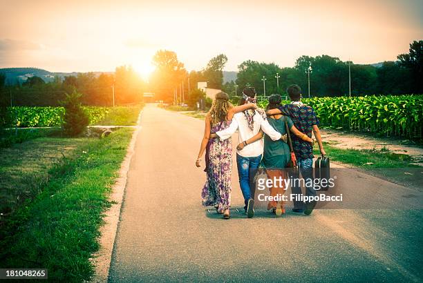 four young adult hippie friends walking on countryside against sunset - arm in arm stock pictures, royalty-free photos & images