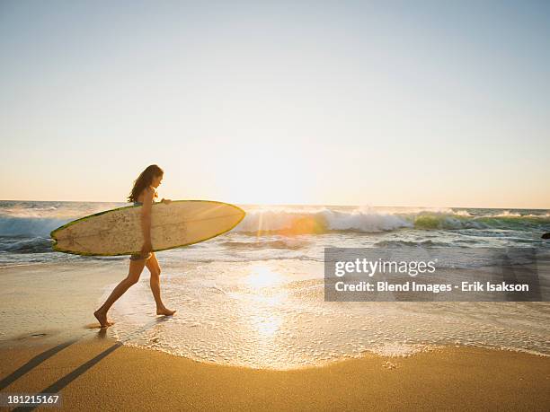 mixed race woman carrying surfboard on beach - californie surf stockfoto's en -beelden