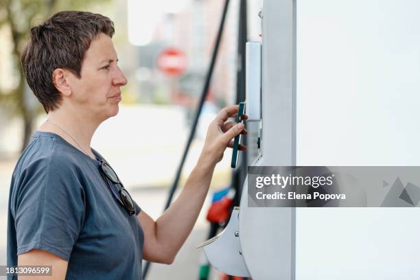 mature adult woman traveling alone, refueling car at self service gas station, paying contactless and looks worried about price for diesel - expense fraud stock pictures, royalty-free photos & images