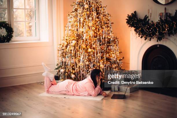 young woman using laptop computer while lying by christmas tree at home - luxury pyjamas stock pictures, royalty-free photos & images