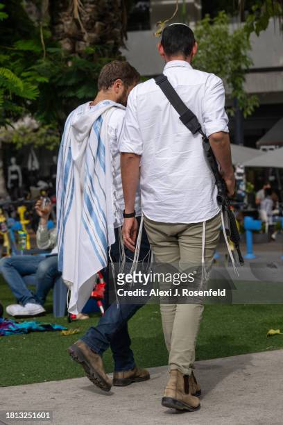 Man wearing a talit walks next to an off duty member of Israeli security forces on Shabbat morning in Dizengoff Square on November 25, 2023 in Tel...