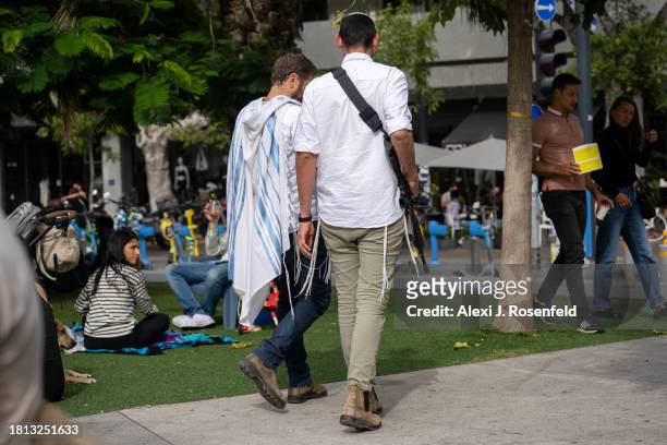 Man wearing a talit walks next to an off duty member of Israeli security forces on Shabbat morning in Dizengoff Square on November 25, 2023 in Tel...
