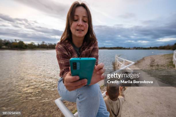 portrait of woman with birthmark on face sitting by lake with smart phone in her hands while her son walking nearby. - modern zagreb stock pictures, royalty-free photos & images