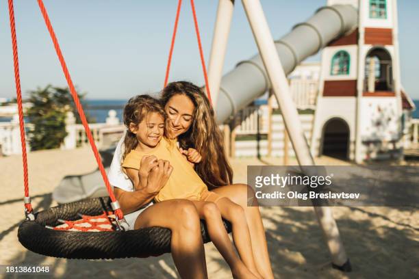 laughing mom swinging with her happy daughter on kids playground. summertime activities - mothers day beach fotografías e imágenes de stock
