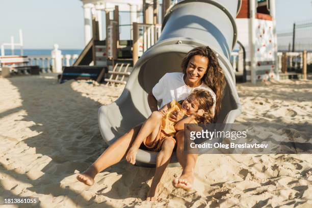 happy mother with her daughter having fun in tube slide on playground. urban environment for children on sandy beach. - mothers day beach fotografías e imágenes de stock