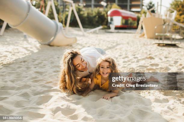 young happy mother playing and having fun with her mischief laughing daughter in sand on beach playground - mothers day beach fotografías e imágenes de stock