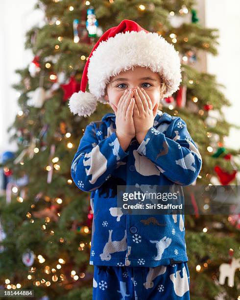 boy wearing santa hat with hands over his mouth - father christmas hat stock pictures, royalty-free photos & images