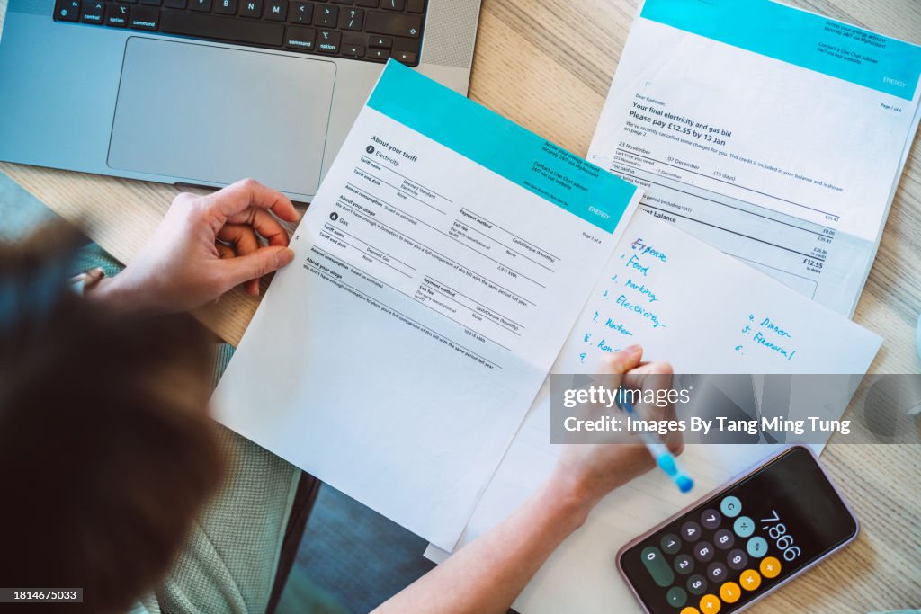 Overhead view of young Asian women managing home finance using laptop & smartphone. She is working with household utility bill and calculating expenses at home.