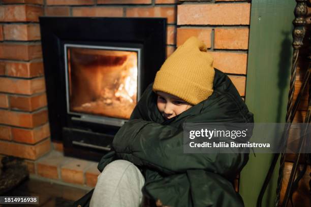 small girl in warm dark jacket sitting beside fireplace in banya feeling cold - wood burning stove fotografías e imágenes de stock