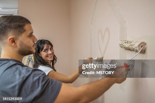 Man painting the wall with paint roller, while his girlfriend writing heart on the wall.