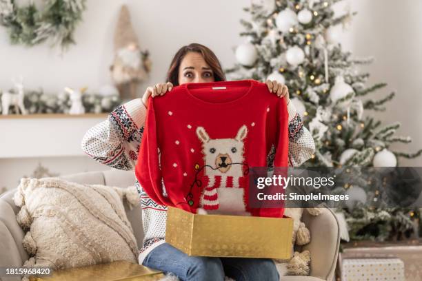disappointed sad woman opening christmas gifts at living room, she has received an ugly and kitsch christmas sweater. - christmas sweater stock pictures, royalty-free photos & images