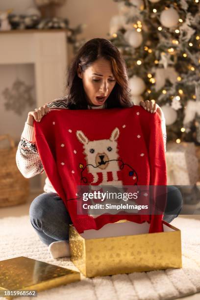 disappointed sad woman opening christmas gifts at living room, she has received an ugly and kitsch christmas sweater. - maglione foto e immagini stock