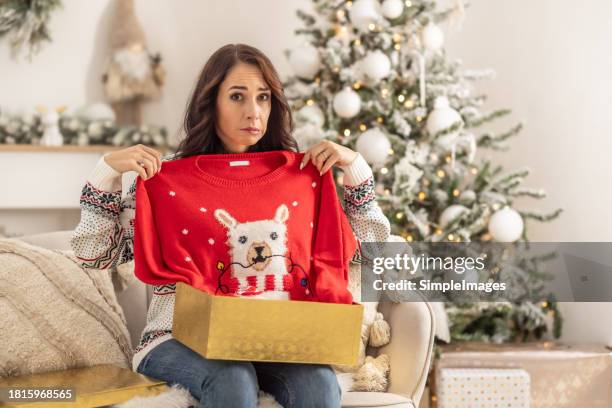 disappointed sad woman opening christmas gifts at living room, she has received an ugly and kitsch christmas sweater. - déception photos et images de collection