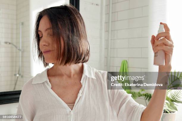 woman spraying cosmetic product on her hair from a bottle while standing in the bathroom - spraying stock pictures, royalty-free photos & images
