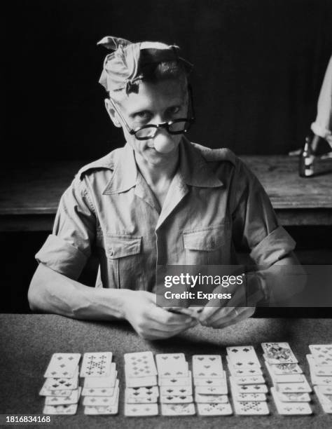 Man wearing a knotted hankie on his head and 'nose glasses', the sleeves of his shirt rolled up as he sits playing solitaire, circa 1955.