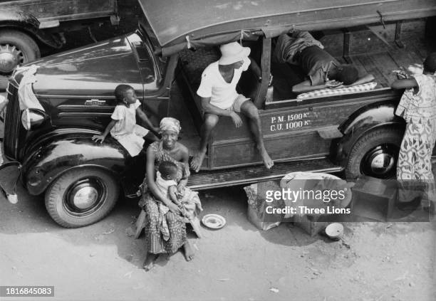 High-angle view of a group of people sitting in, on and around a Chevrolet vehicle at a transport company in Kpalime, Togo, circa 1955. The woman...