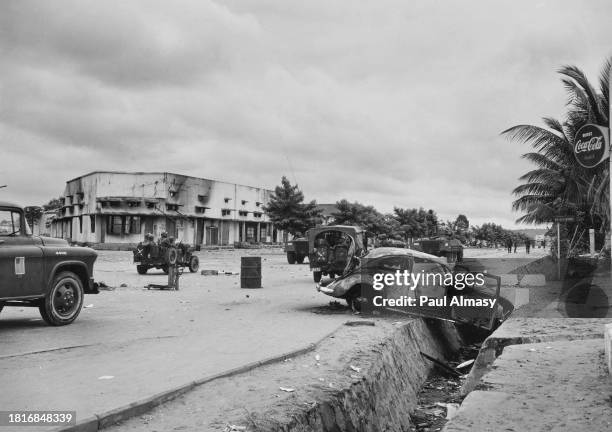 Military personnel pass along an otherwise deserted street, with burnt-out vehicles following rioting in Leopoldville, Belgian Congo, January 1959....