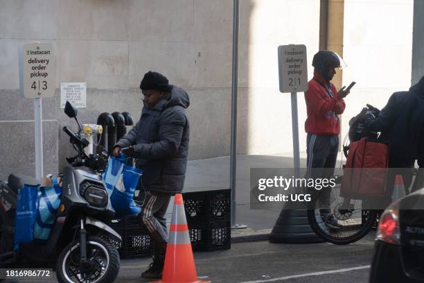 Delivery worker balances a bag against his motorbike while simultaneously looking down at his phone, another checks his phone, and a third stands by...