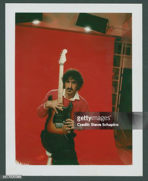 Portrait of American musician Frank Zappa as he poses, with a guitar, in front of a red background, Los Angeles, California, 1984.