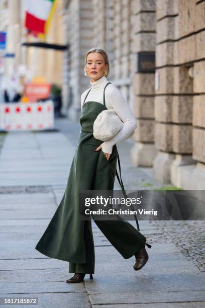 Tina Haase wears Karpova green wool pants & apron dress, white COS Teddy Clutch bag, white turtleneck, Zara heeled boots on November 27, 2023 in...