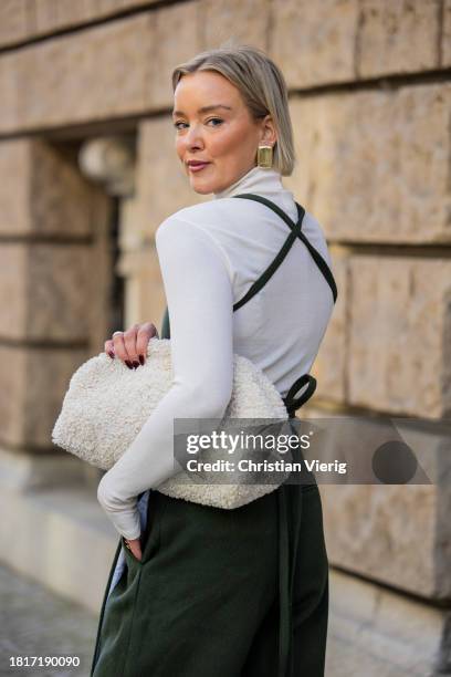 Tina Haase wears Karpova green wool pants & apron dress, white COS Teddy Clutch bag, white turtleneck, Zara heeled boots on November 27, 2023 in...