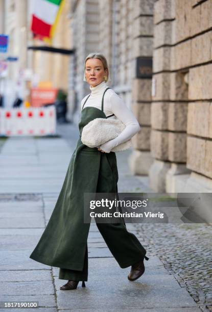 Tina Haase wears Karpova green wool pants & apron dress, white COS Teddy Clutch bag, white turtleneck, Zara heeled boots on November 27, 2023 in...