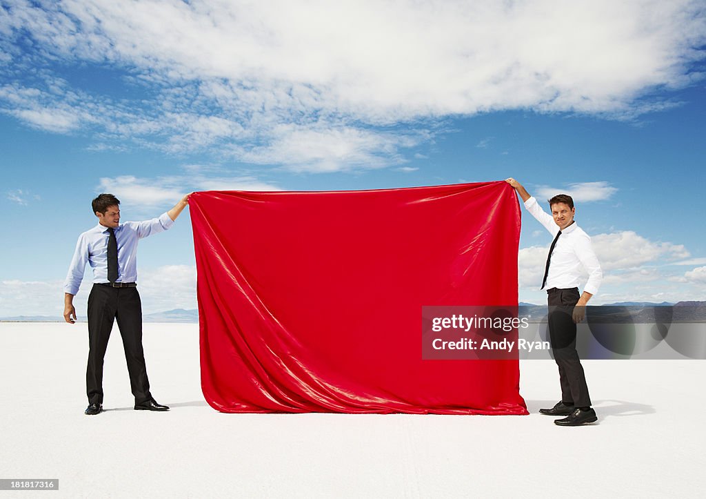 Two men holding up sheet of red fabric in desert.