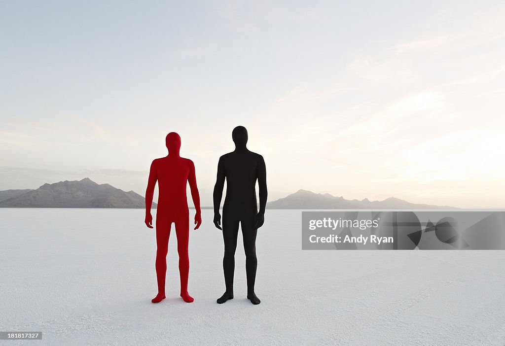 Two men in bodysuits stand in desert.