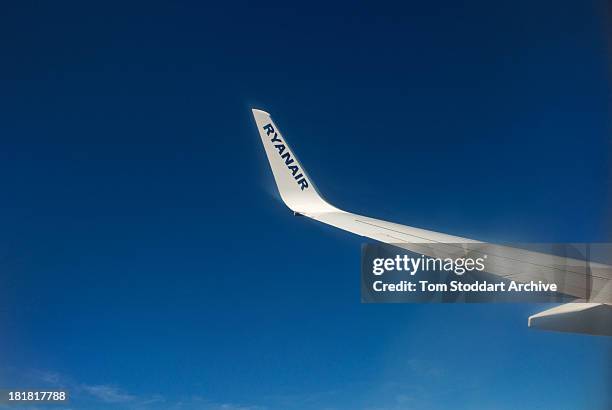 Photograph of a wing of a Ryanair plane flying in the blue sky towards Stansted Airport in Essex, England. Ryanair was founded in 1985 in Dublin,...