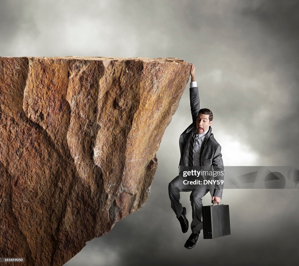 Businessman hanging on for dear life from edge of cliff