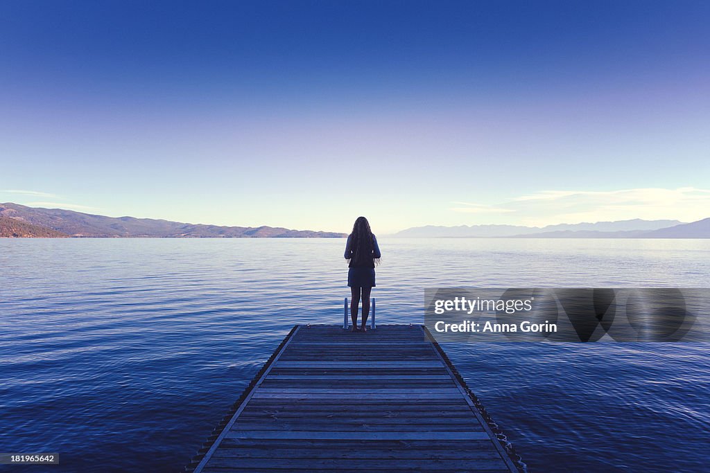 Silhouetted woman stands at edge of dock on lake