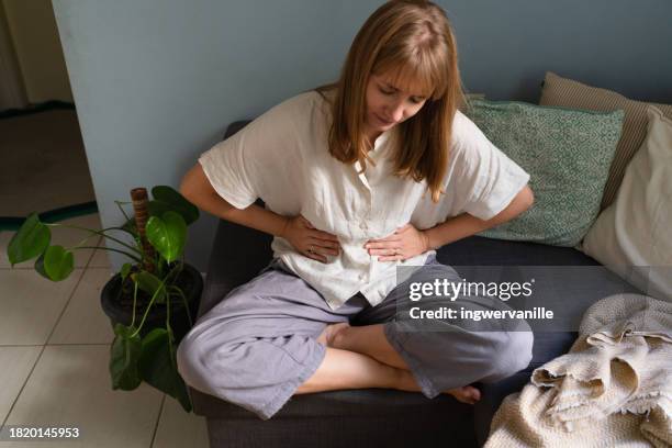woman sitting on sofa and holding hands on stomach. pain - brechreiz stock-fotos und bilder