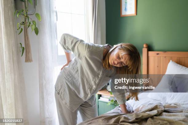 grimacing woman suffering from pain in back leaning against bed - backache stock pictures, royalty-free photos & images