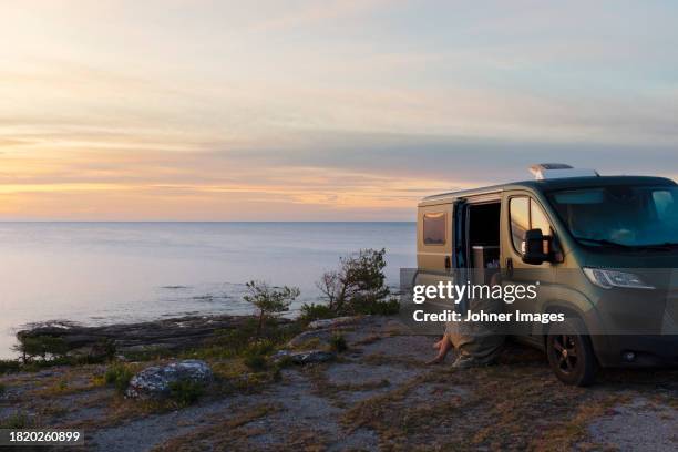 woman sitting near motorhome and looking at sea - lieferwagen stock-fotos und bilder