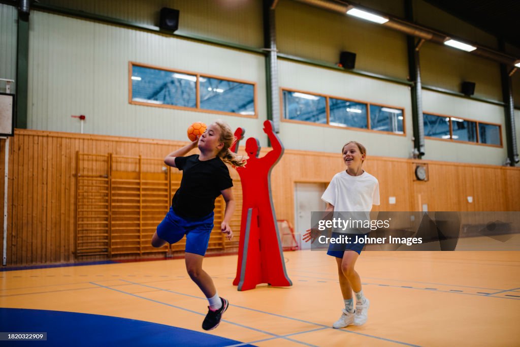 Girl jumping while playing handball with female friend in sports court