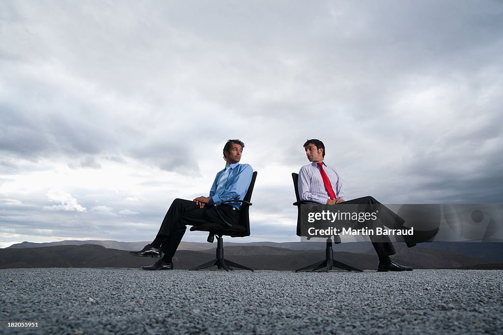 Two men sitting in office chairs outdoors with their backs against one another