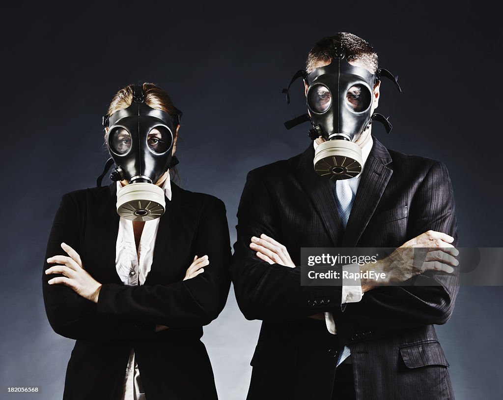 Formally dressed couple in gas masks fold arms and stare