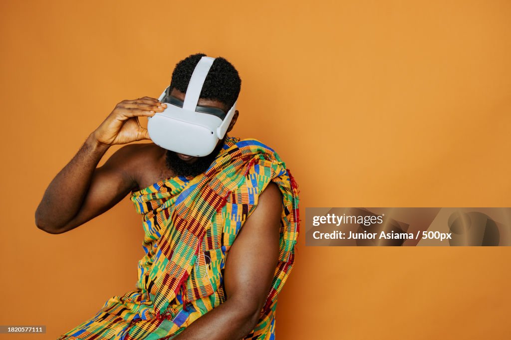 Man wearing virtual reality simulator while wearing traditional clothing against orange background,Accra,Ghana