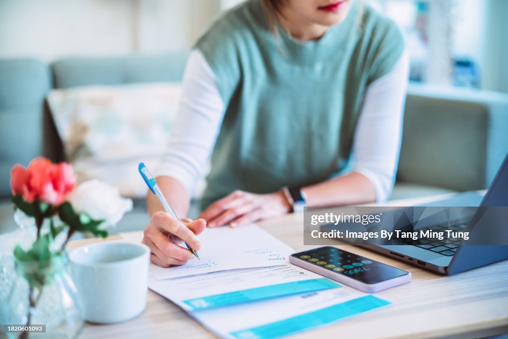 Young Asian women managing home finance using laptop & smartphone. She is working with household utility bill and calculating expenses at home.