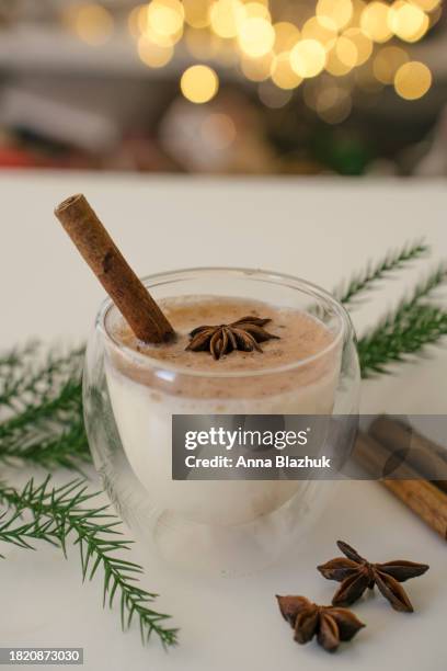 traditional christmas dring eggnog with cinnamon in glass on white table. festive setting, green pine tree and golden blurred lights in background. - eggnog stock pictures, royalty-free photos & images