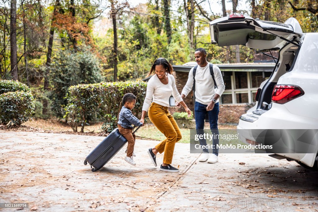 Mother pulling young boy on suitcase and loading car for family vacation