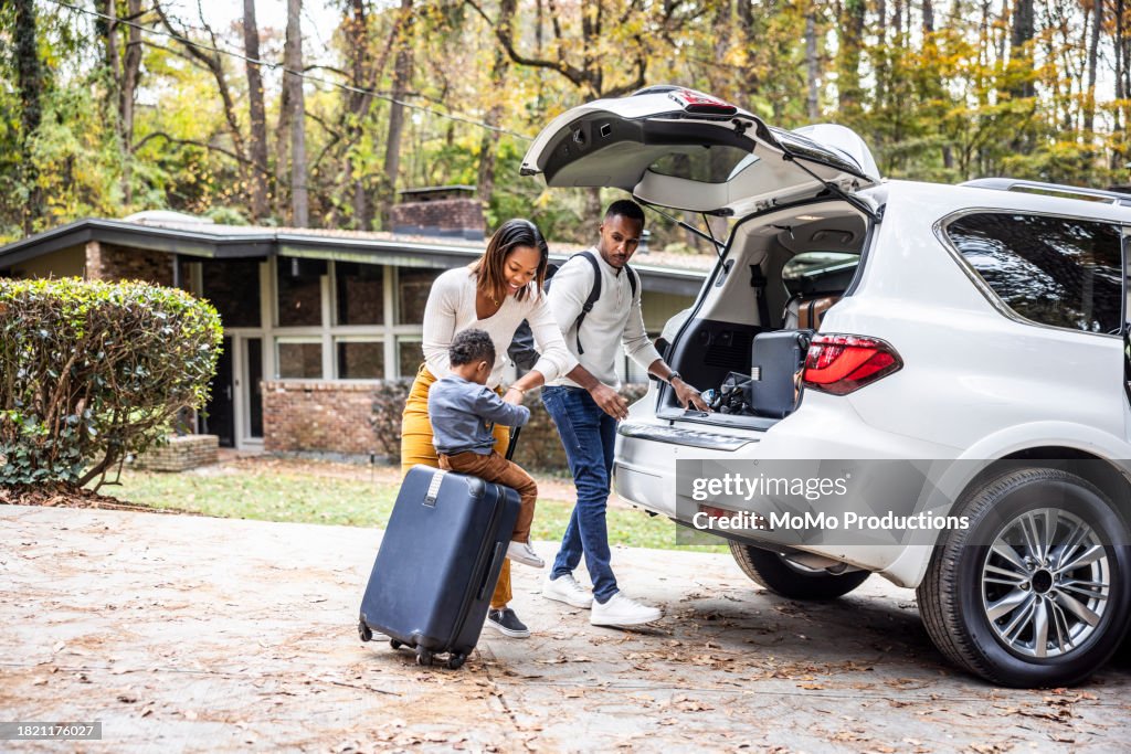 Mother pulling young boy on suitcase and loading car for family vacation