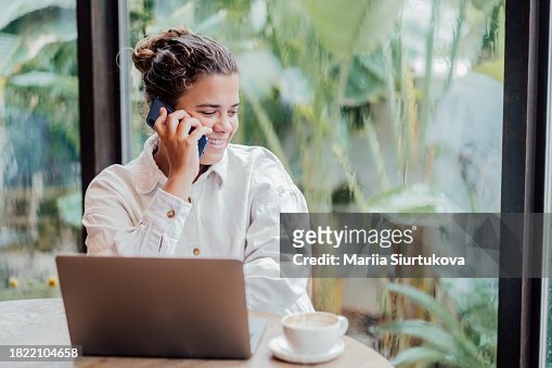 Young woman chats on mobile phone while working at laptop computer near large panoramic window.