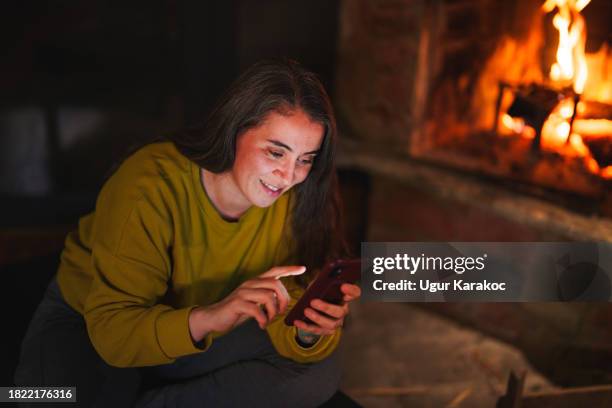 woman using mobile phone in front of a fire place at home - comfort zone stock pictures, royalty-free photos & images