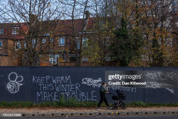 Member of the public walks past a graffiti sign that reads 'Keep warm this winter, make trouble' on November 30, 2023 in Harrow, London, United...