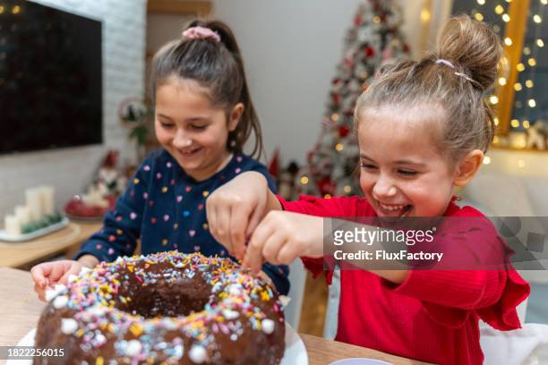 adorable caucasian sisters decorating the chocolate bundt cake together with sprinkles - tulbandcake stockfoto's en -beelden
