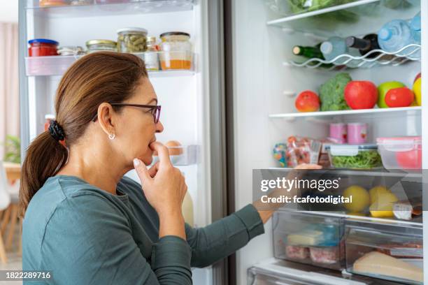 retrato de una mujer dubitativa mirando dentro de la nevera - hambre fotografías e imágenes de stock